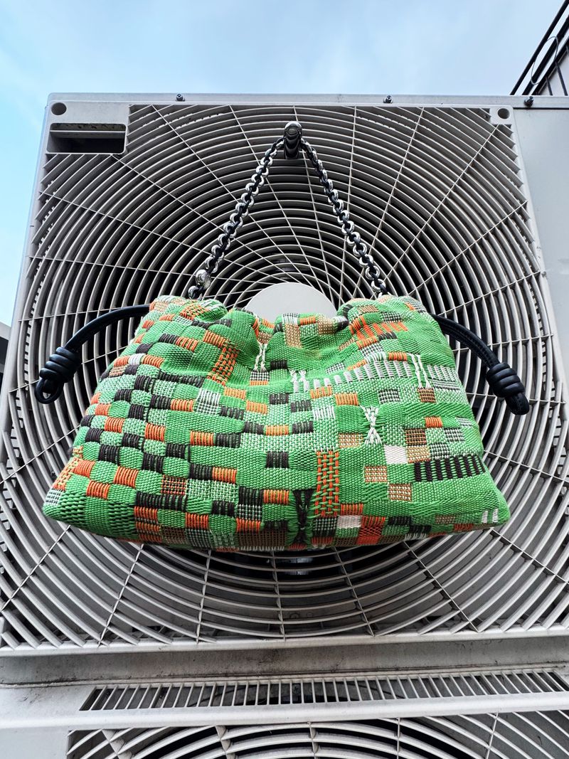 Green woven tote bag with drawstring closure and silver chain straps hanging from a gray ventilation unit against a light blue sky.