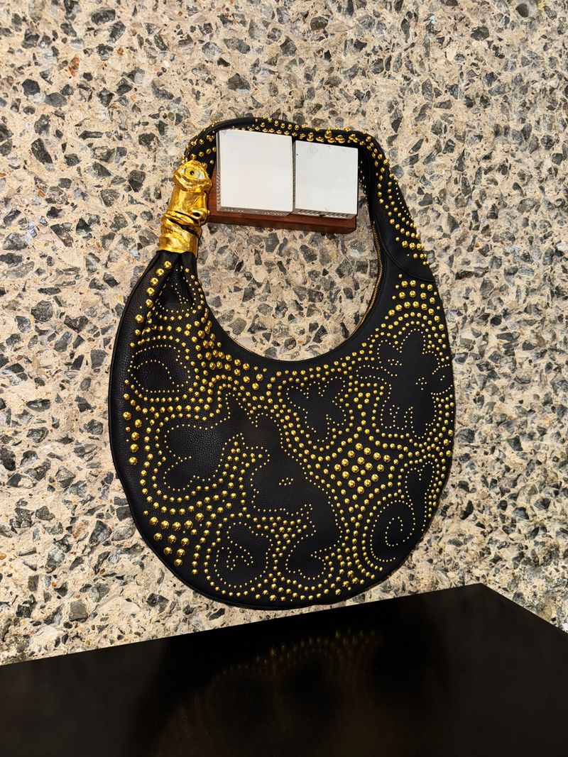 Close-up of a stylish black leather handbag embellished with gold studs, featuring a unique gold handle on a patterned surface.