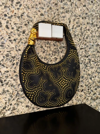 Close-up of a stylish black leather handbag embellished with gold studs, featuring a unique gold handle on a patterned surface.