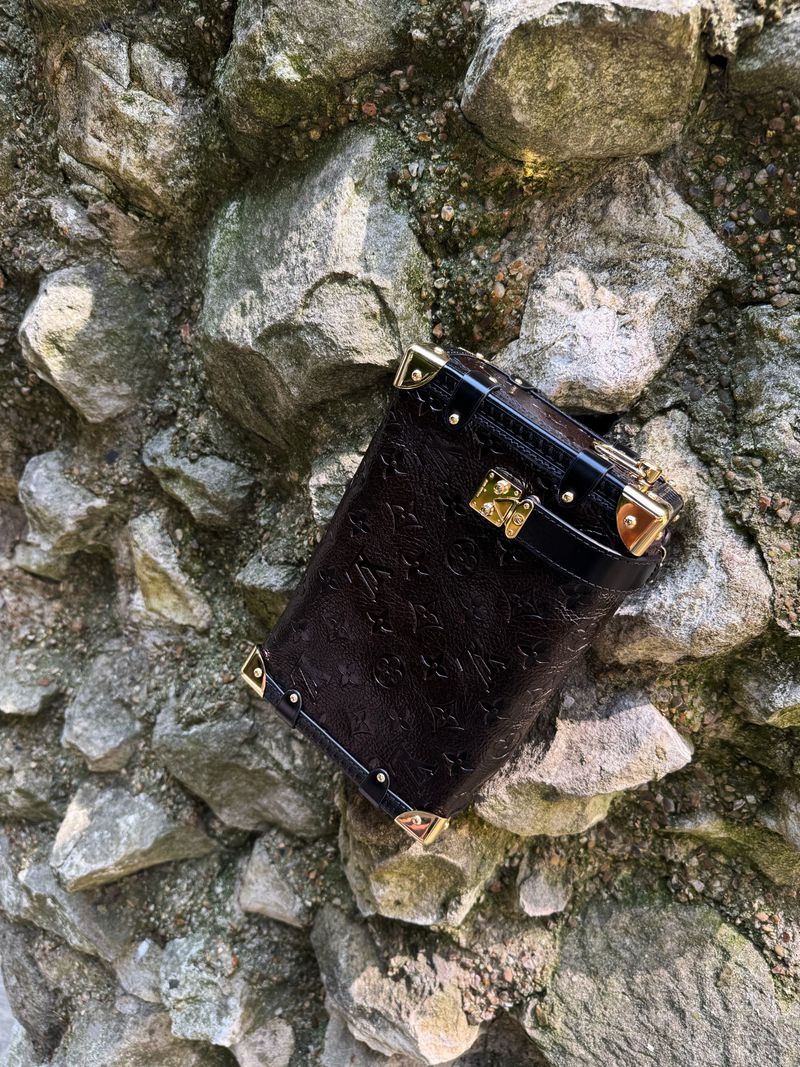 Elevated shot of a small, black Louis Vuitton trunk with gold accents, set against a textured, light-grey rock background.