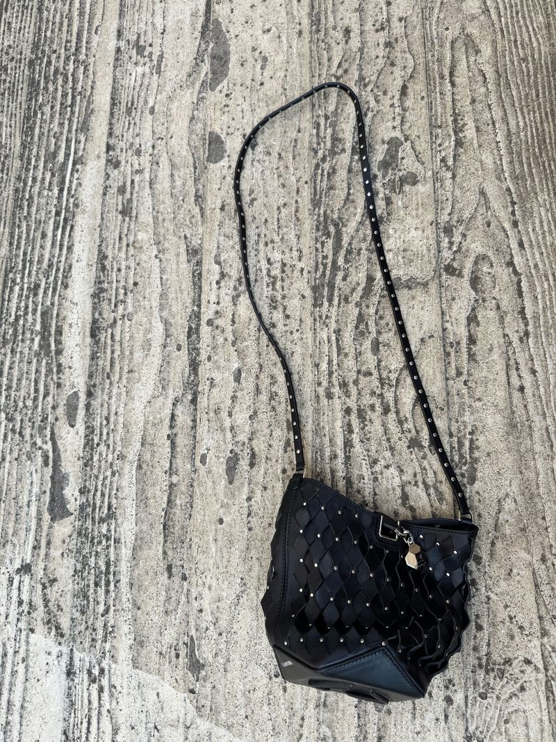 Black woven handbag with a shoulder strap is displayed against a textured gray and white wood-look backdrop in a product-style shot.