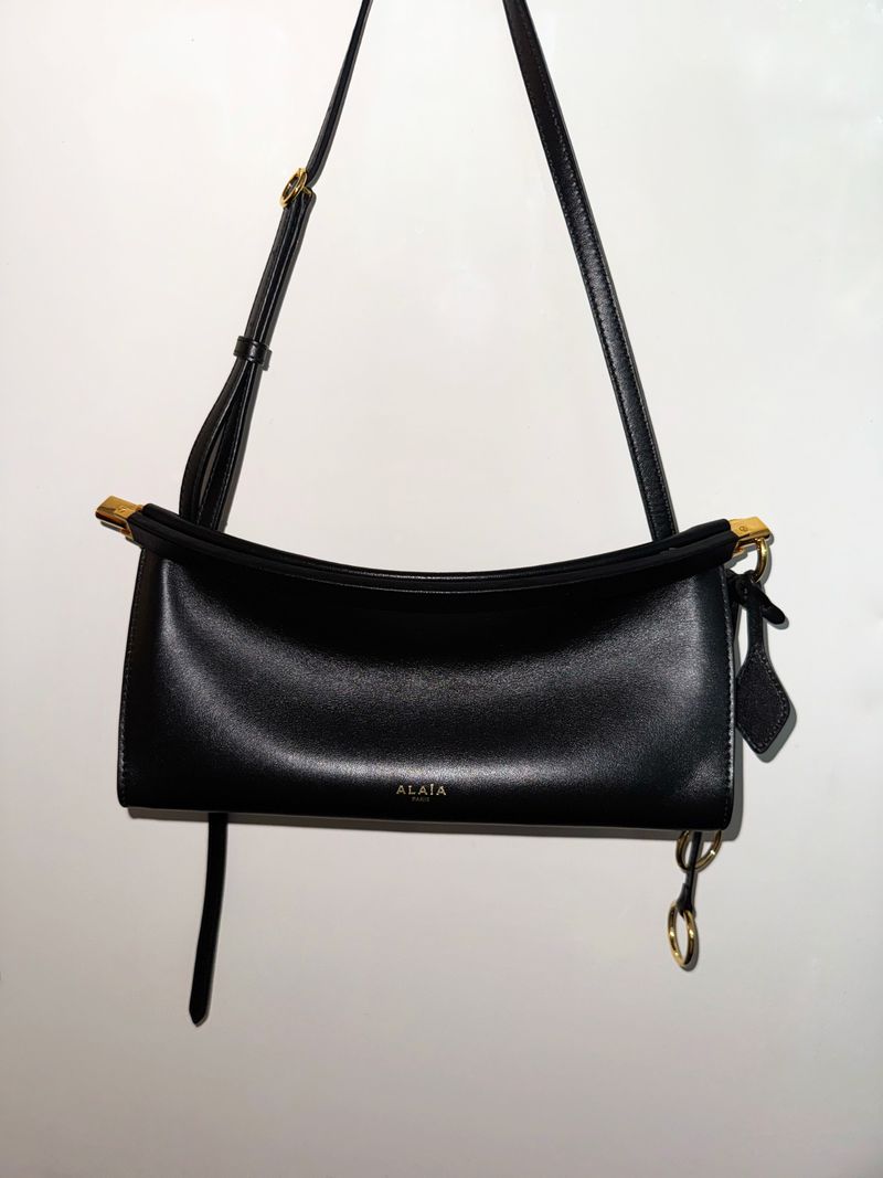 Studio shot of a black leather Alaïa handbag with a gold-tone hardware strap, hanging against a white background. Fashion product.