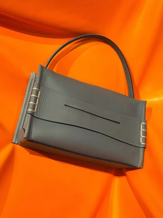 Gray leather shoulder bag with gold hardware against a vibrant orange backdrop, highlighting texture and design in a fashion product shot.