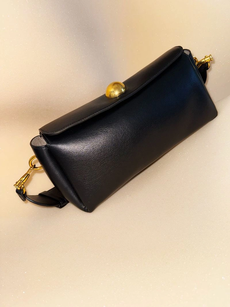Black leather clutch handbag with a round gold sphere clasp resting diagonally on a shimmering beige surface.