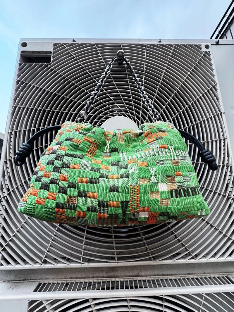 Woven green and orange patterned handbag hanging from a silver chain strap on an industrial air conditioning unit fan grille.