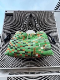 Woven green and orange patterned handbag hanging from a silver chain strap on an industrial air conditioning unit fan grille.