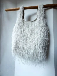 White fringed textured tote bag hanging from a natural wood rail against a pale wall.