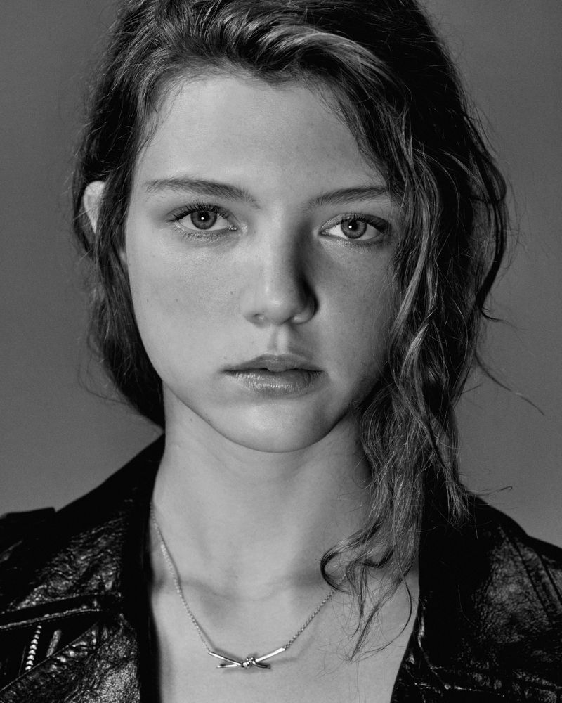 Black and white portrait of a young woman with wavy brown hair wearing a black leather jacket and a silver necklace, shot in a photo studio.