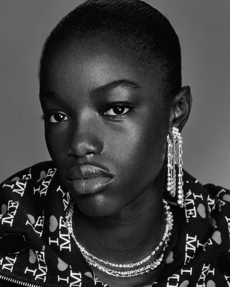 Headshot of a Black woman wearing statement jewelry, including earrings and a necklace, with monochrome lighting in a studio setting.
