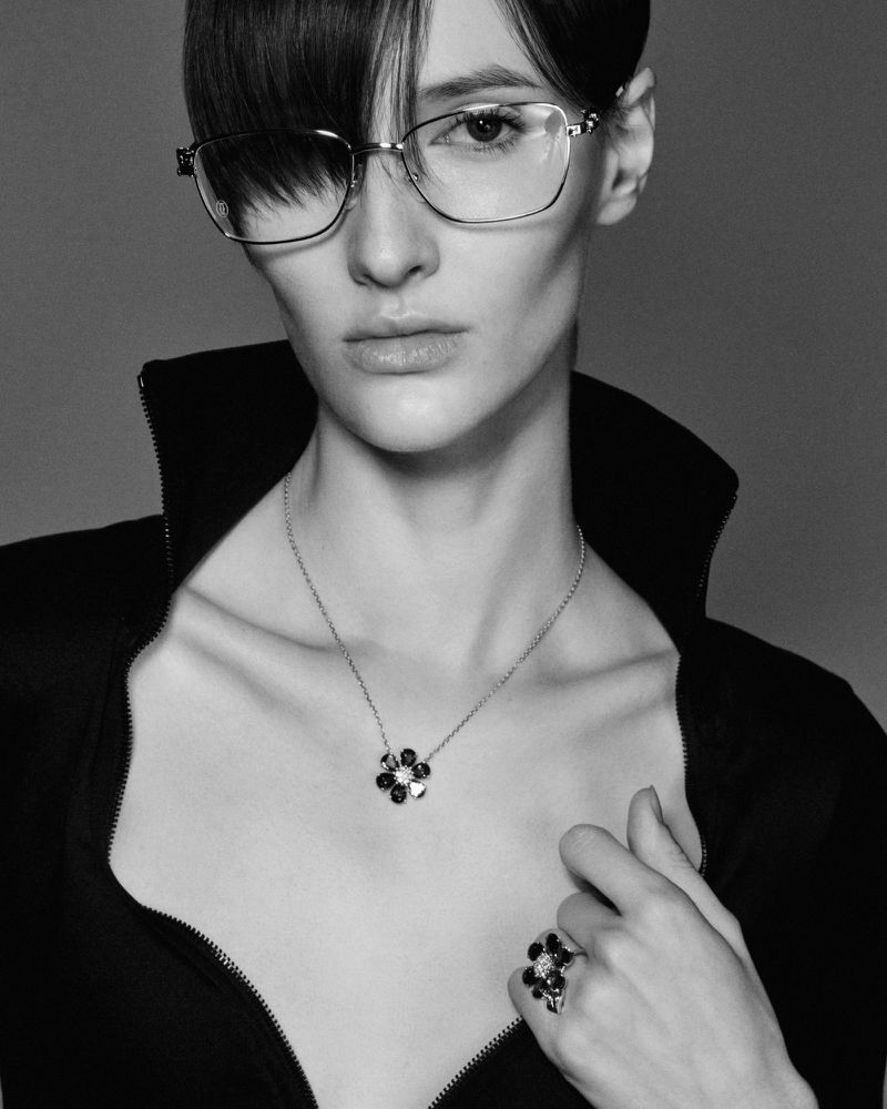 Monochrome portrait of an adult woman with eyeglasses, wearing a black top and floral jewelry, set against a neutral gray backdrop.
