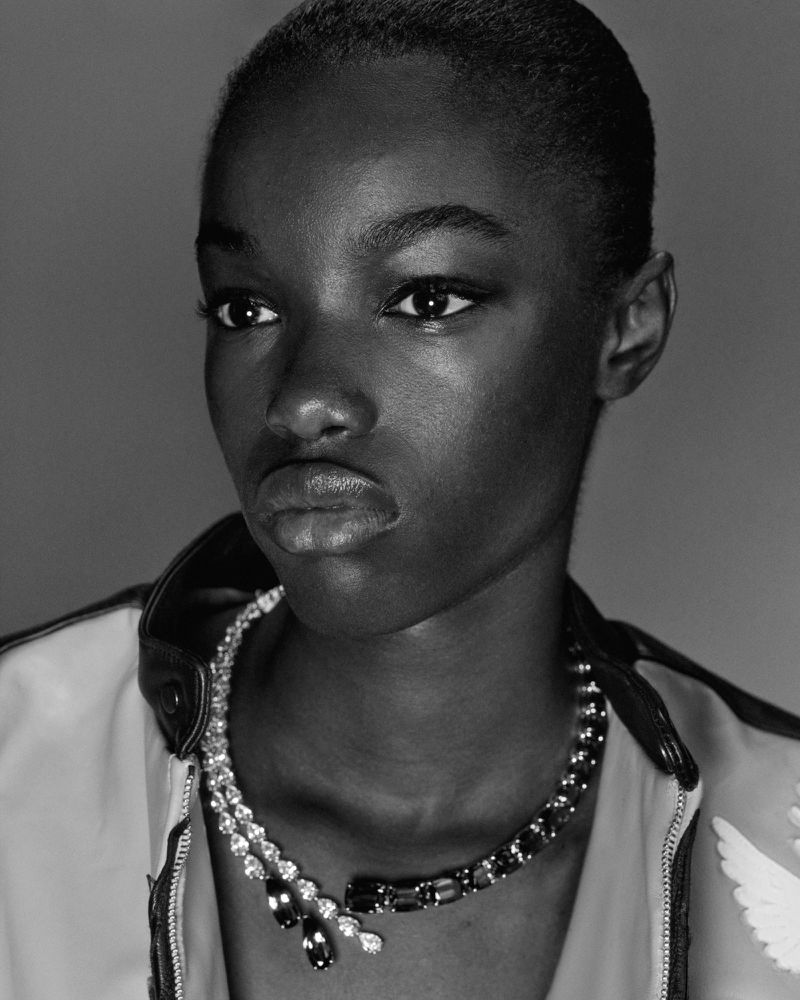 Portrait of a young adult black woman wearing a necklace and jacket against a neutral gray backdrop in a black and white photograph.