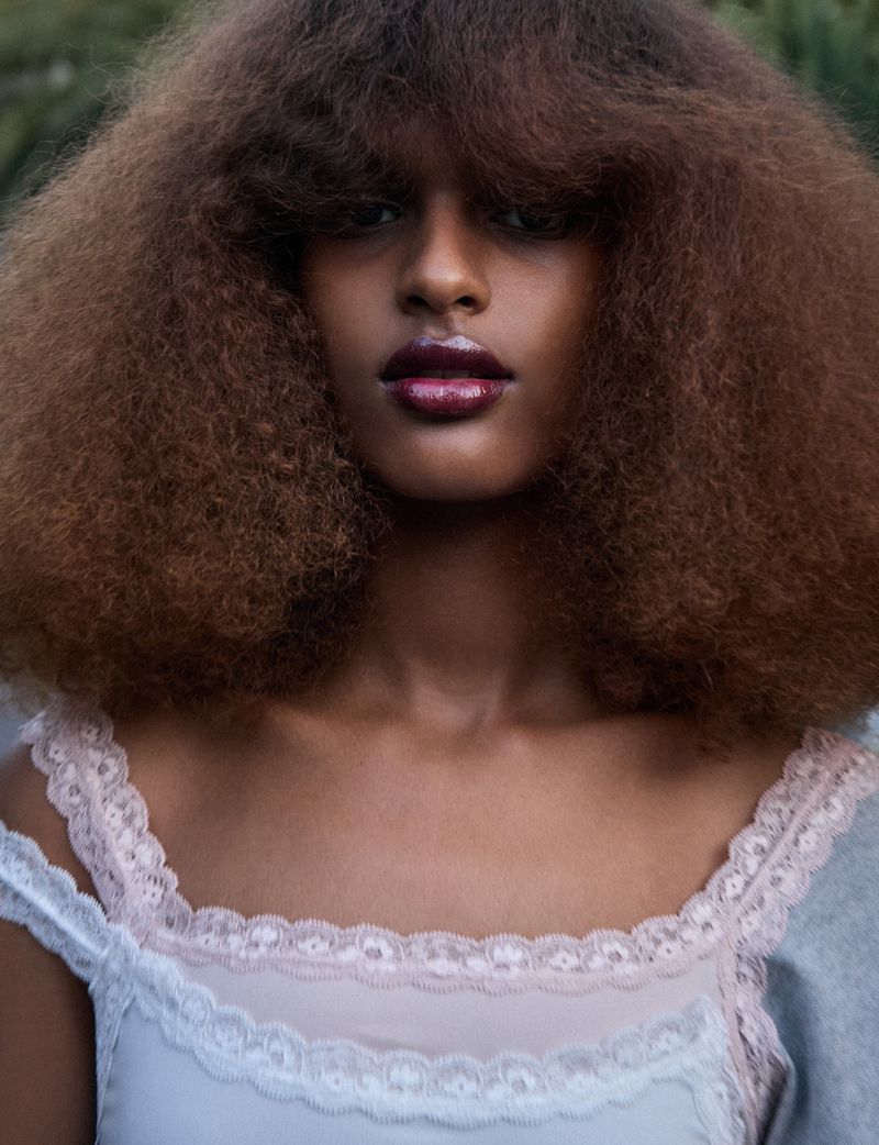 Close-up portrait of a woman with a large afro and light blue and pink lace-trimmed top against a soft green outdoor background.