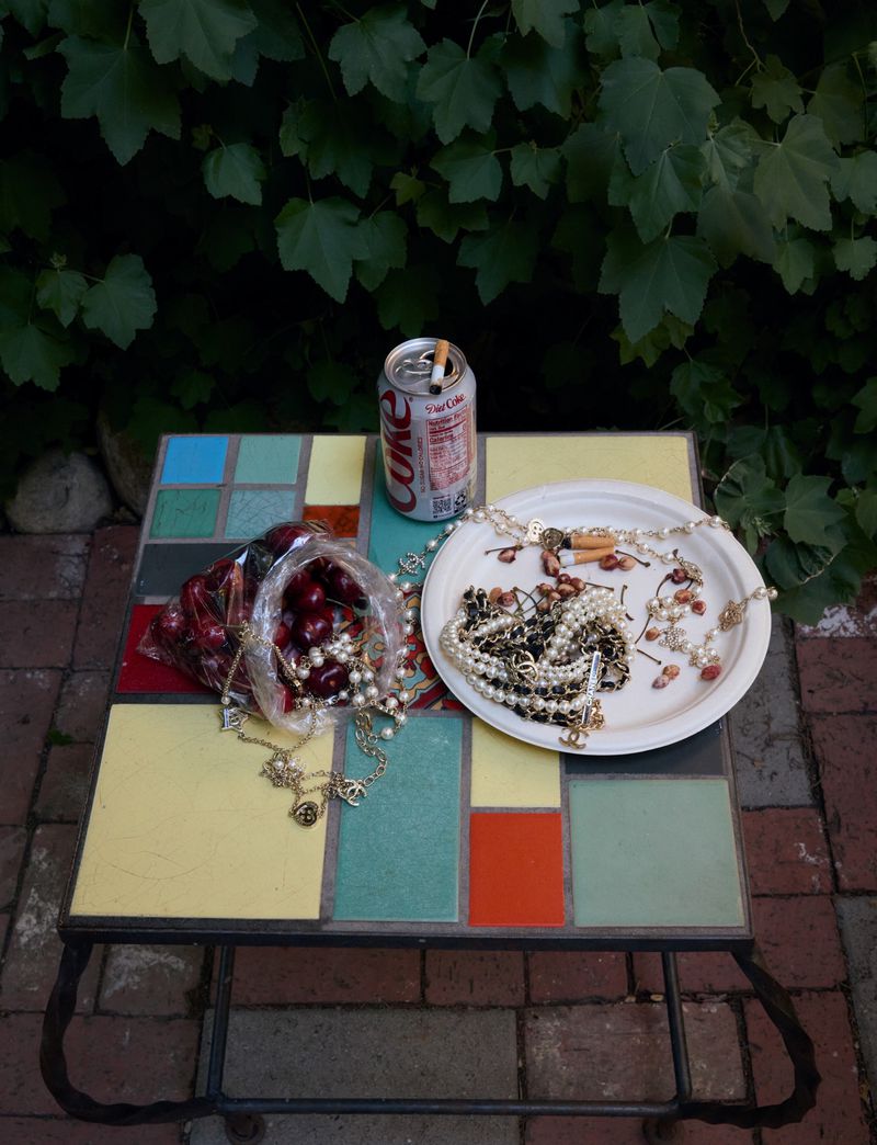 Still life with a bag of cherries, costume jewelry, and an open can of Coke on a colorful tile-top table outdoors with a green foliage background.