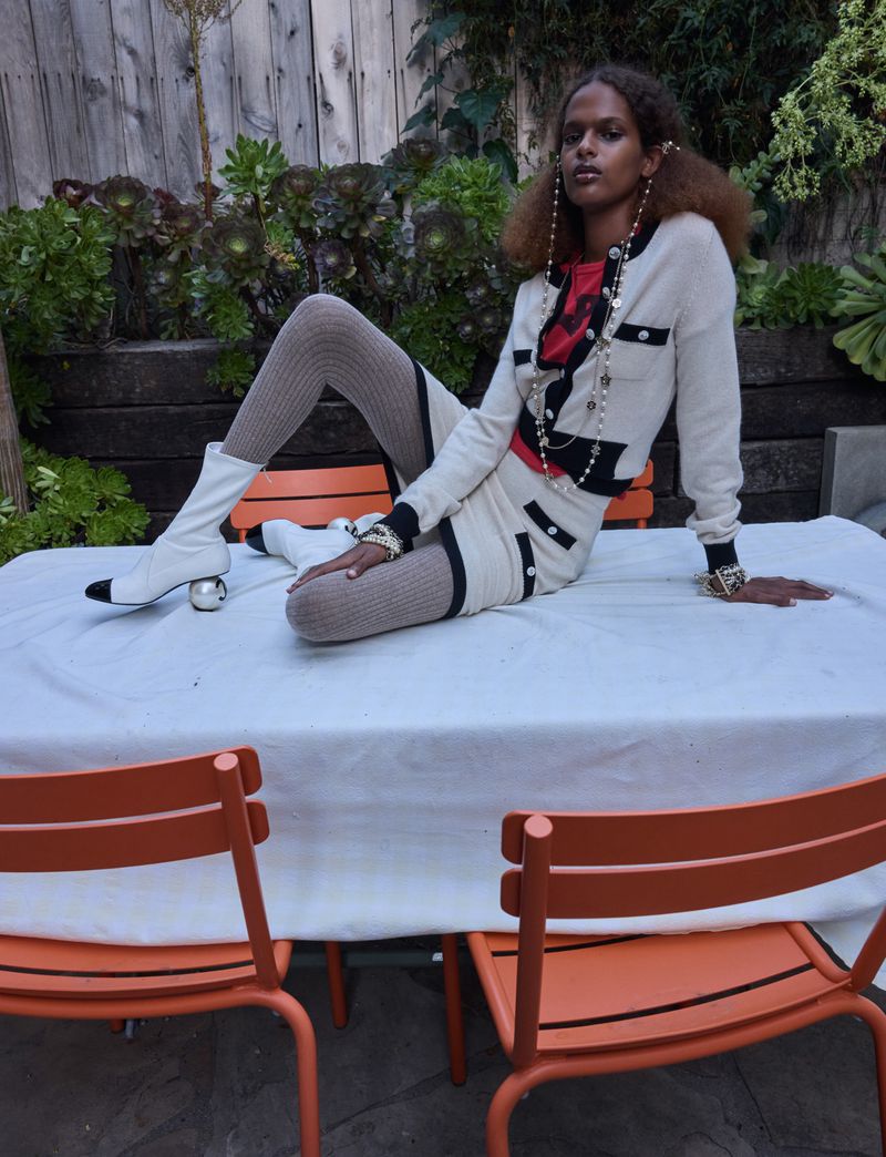Elegant adult female poses on an outdoor table in a fashion editorial shoot, wearing a cream-colored skirt suit and pearl jewelry.