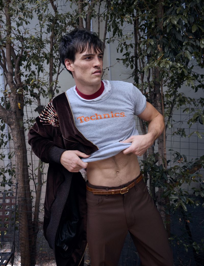 A young adult man stands outdoors lifting his t-shirt slightly to show his abs, wearing brown pants and a brown jacket.
