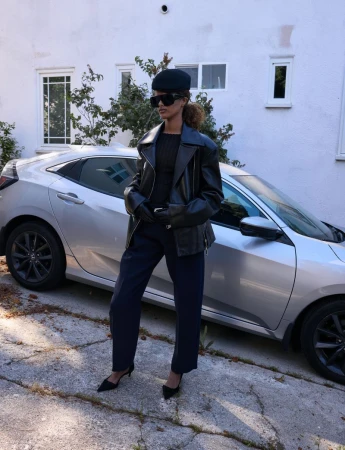 Woman in black jacket and beret poses with a silver car in urban setting, wearing sunglasses and gloves on sunny day.
