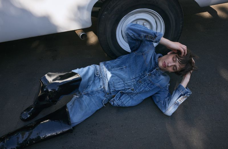 A young adult lying on asphalt in a denim jacket and jeans, wearing black shiny boots, next to the tire of a white vehicle outdoors.