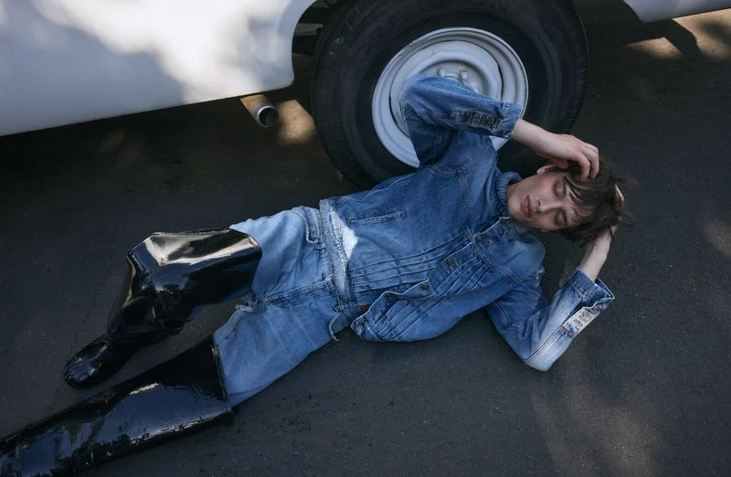 A young adult lying on asphalt in a denim jacket and jeans, wearing black shiny boots, next to the tire of a white vehicle outdoors.
