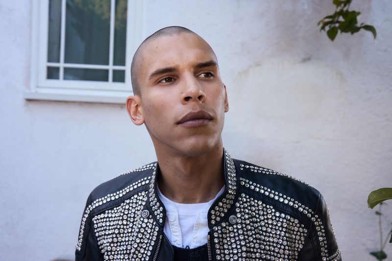 Adult male with a shaved head wearing a black leather jacket with silver studs, photographed outdoors against a white wall and green foliage.