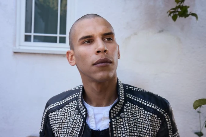 Adult male with a shaved head wearing a black leather jacket with silver studs, photographed outdoors against a white wall and green foliage.