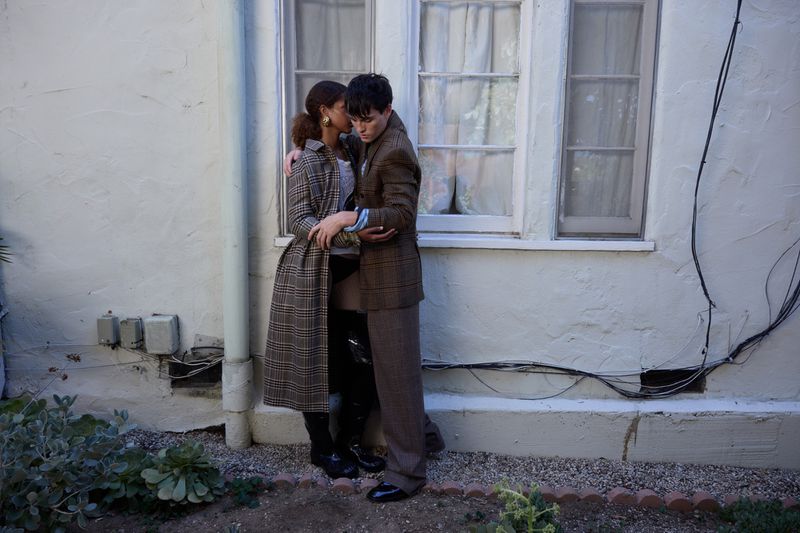 A stylish interracial couple in tailored outfits embraces outdoors near a window, showing fashion and connection in natural light.