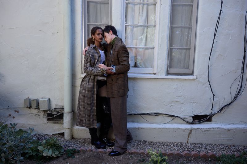 A couple poses in front of a white stucco wall and window, embracing each other, wearing dark colors and earth-toned plaid patterns.