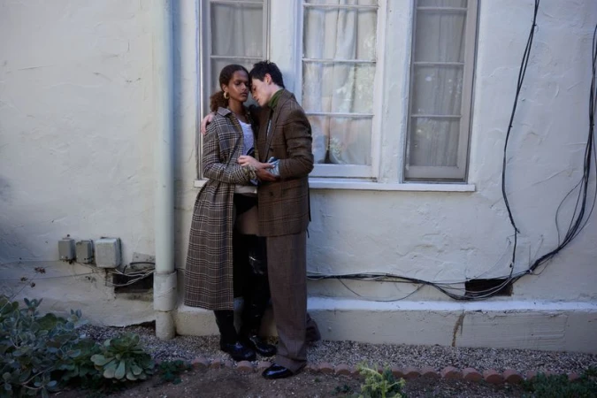 A couple poses in front of a white stucco wall and window, embracing each other, wearing dark colors and earth-toned plaid patterns.