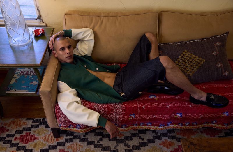 Adult male laying on a couch wearing a green jacket, denim shorts, and black loafers in an indoor environment with patterned rugs.