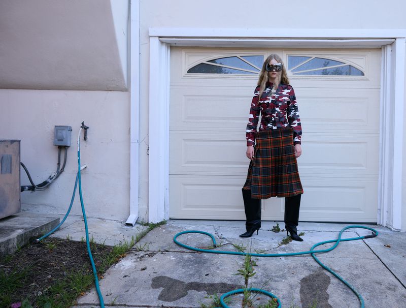 Adult woman in masquerade mask, camouflage top, and plaid skirt poses on a cracked driveway in front of a closed garage door.