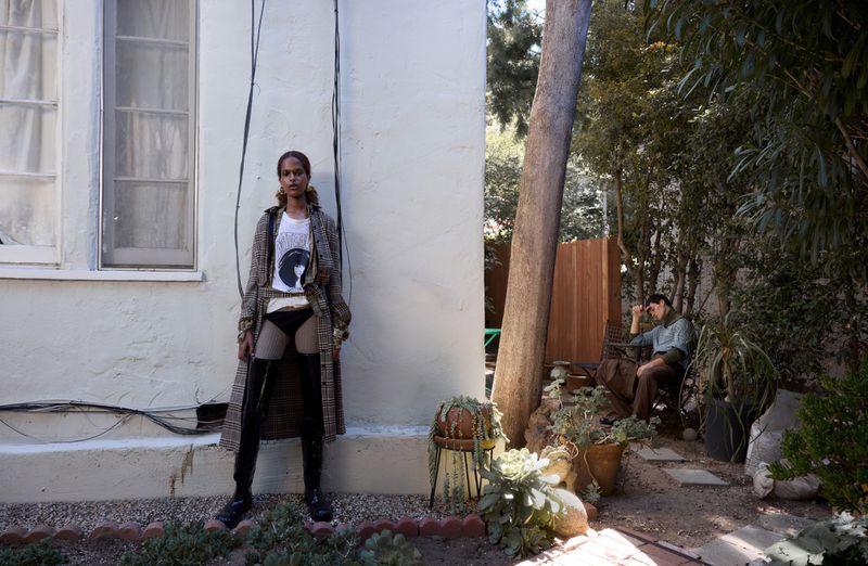 Woman in plaid coat and thigh-high boots poses against a white wall in an outdoor setting; man sits in background surrounded by lush greenery.
