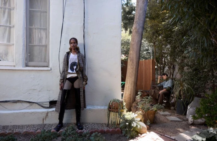 Woman in plaid coat and thigh-high boots poses against a white wall in an outdoor setting; man sits in background surrounded by lush greenery.