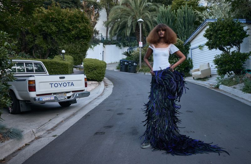 Woman with large afro hairstyle poses in a feathered skirt on a street next to a parked Toyota pickup truck in daylight.