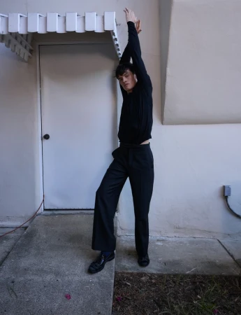 A slender young adult male model posing outside a building in a black sweater, dress pants, and patent-leather dress shoes in a fashion editorial.