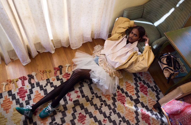 Woman in yellow jacket and tutu reclines on patterned rug in sunlit room near armchair and side table in top-down fashion photograph.