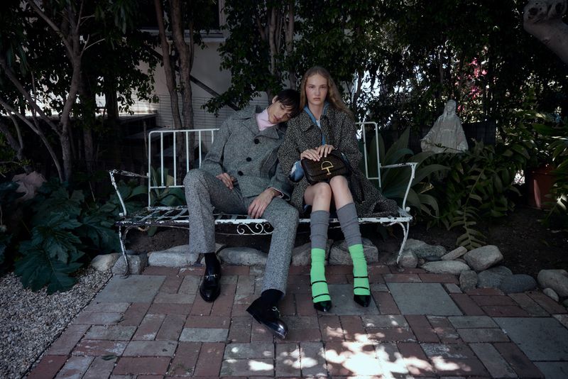 A young couple poses on a white iron bench in a garden setting, wearing patterned grey outerwear and black footwear.  