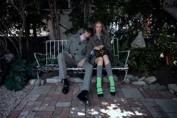 A young couple poses on a white iron bench in a garden setting, wearing patterned grey outerwear and black footwear.  