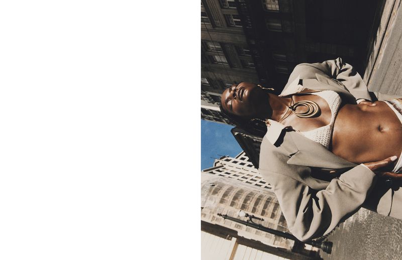 Woman in blazer and knit bra poses in an urban outdoor setting with high-rise buildings, shot from a low angle under natural sunlight.
