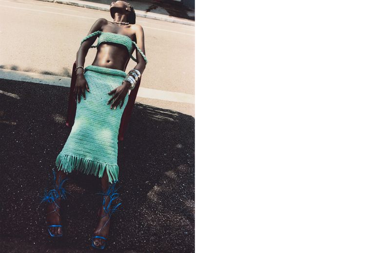An adult woman in an outdoor fashion shot lies on a road in a light green crochet skirt, top, and blue feathered high heels, sunny.
