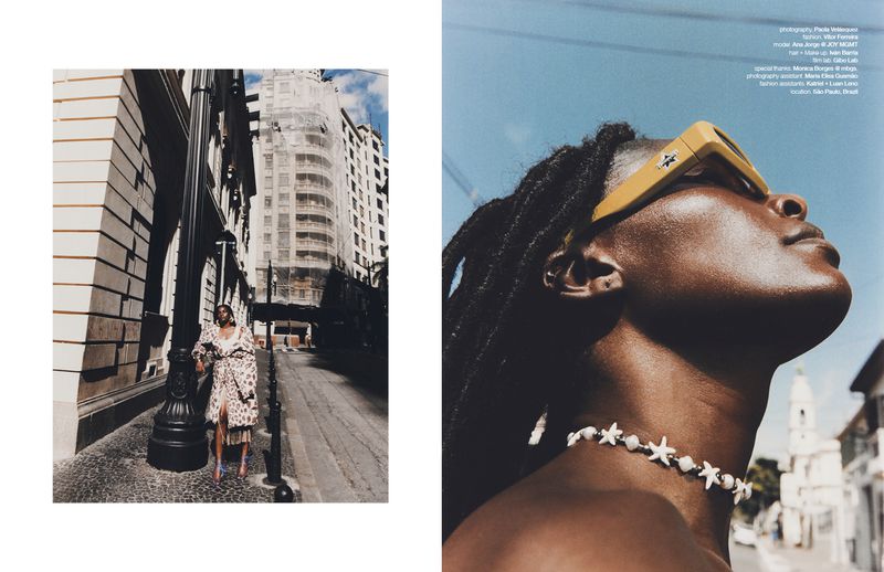 Fashion editorial shot with an adult female model posing outdoors in a coat, buildings in the background, sunglasses and shell necklace.
