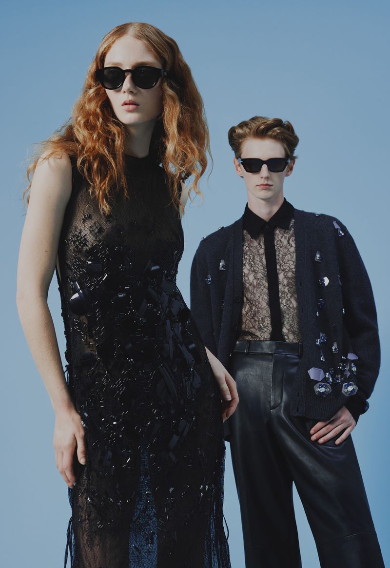 Two fashion models wearing sunglasses and designer clothing pose against a light blue backdrop in a studio shot with editorial style.