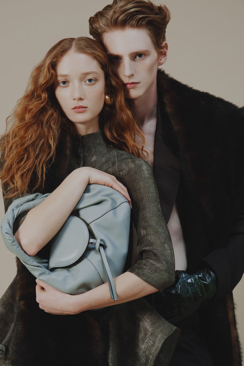 Fashion shot featuring a young couple; a woman holding a light blue handbag while posing with a man in a fur-trimmed coat in a studio.
