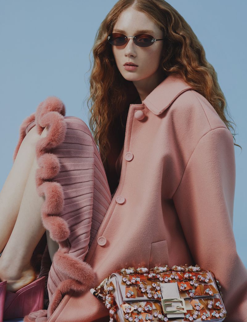 Fashion photography of a woman wearing sunglasses, a pink coat, and a patterned purse against a light blue backdrop in a studio.