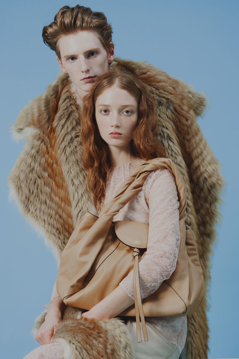 Fashion shot featuring two models, one male and one female, with matching red hair. The female model is holding a beige handbag.