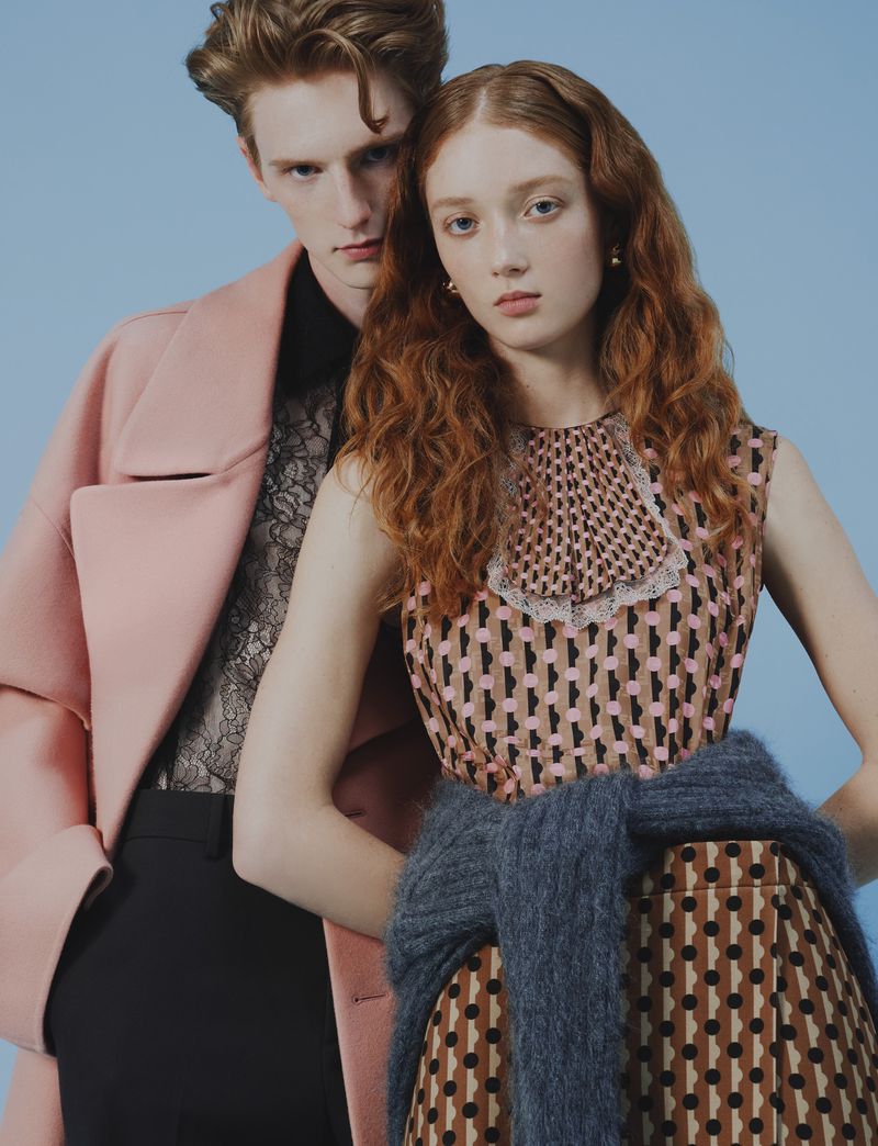 A stylish fashion portrait of a red-haired woman in a patterned dress and a man in a pink coat against a light blue background.