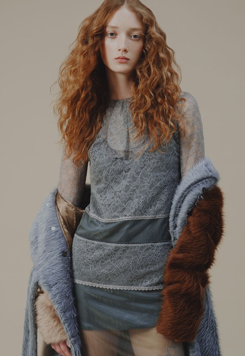 Elegant redhead woman in blue lace top and long fur coat poses in a fashion studio with soft, diffused lighting and a neutral backdrop.