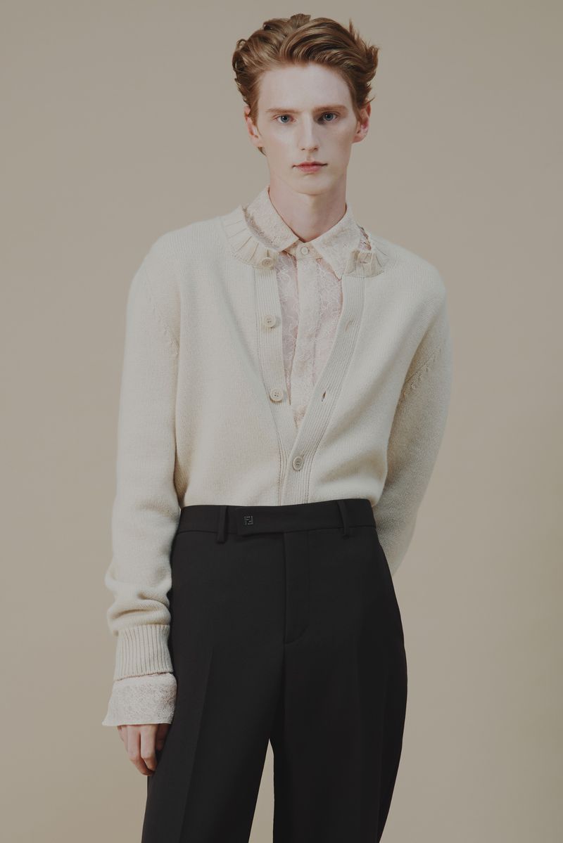 Pale ginger man in a buttoned cream cardigan over patterned shirt with black pants poses indoors against a plain neutral backdrop.