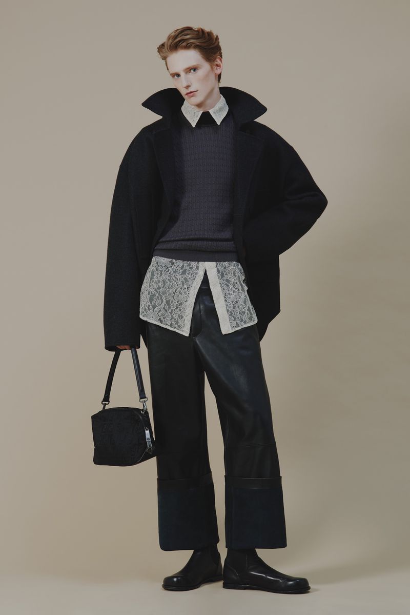 Studio shot of a young adult man wearing a black coat and dark denim pants, holding a black handbag, posing against a beige background.