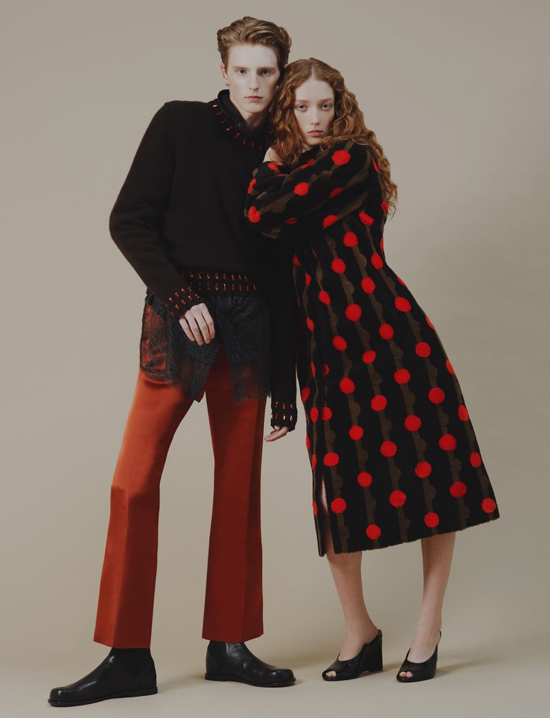 Two models pose in a studio, one wearing a black sweater and rust-colored pants, the other in a red and black polka-dot dress.