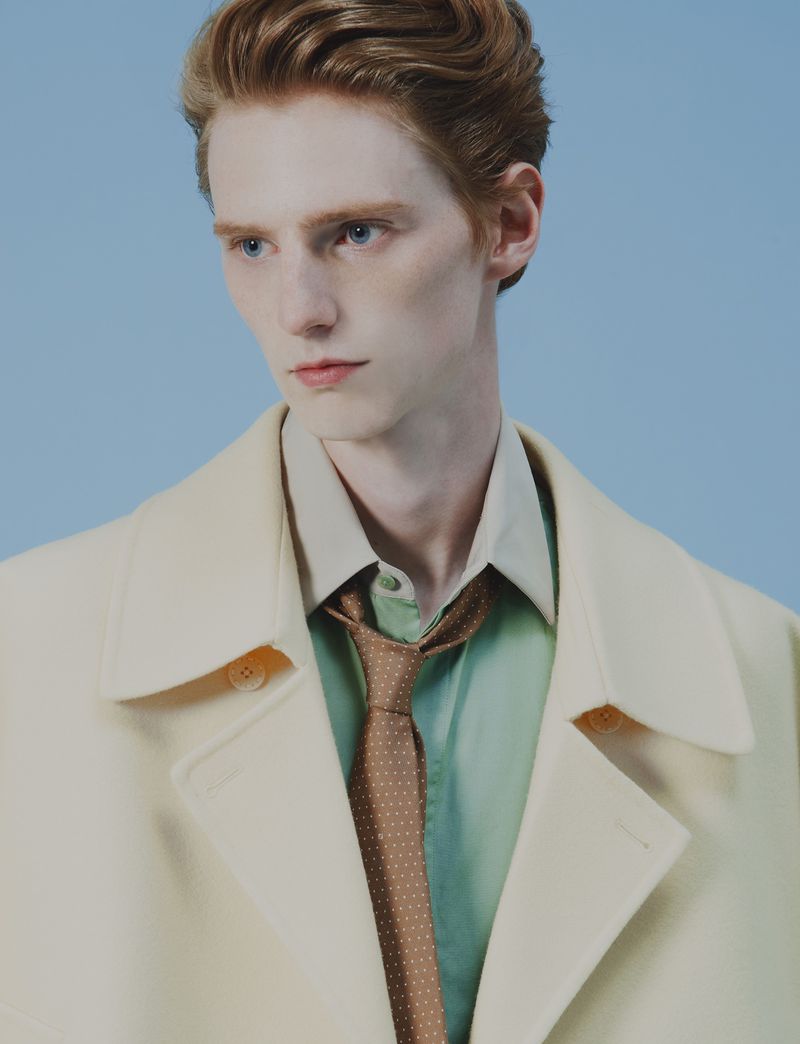 Man in beige coat and tie against blue backdrop. Ginger hair and blue eyes. Classic menswear. Fashion shot, portrait orientation.