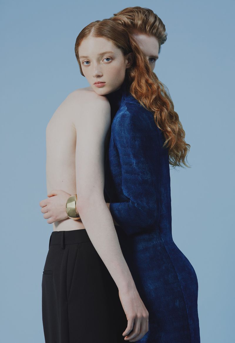 Studio shot of two red-haired models embracing, one with bare chest and gold bangle, the other in a blue, textured dress, against blue backdrop.